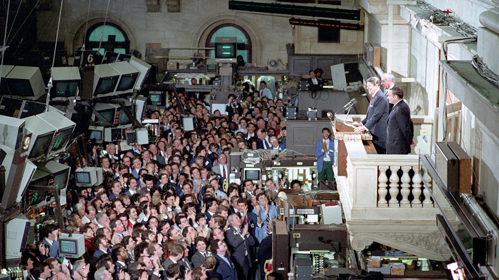 A crowded New York Stock Exchange trading floor in the 1980s, packed with brokers and rows of monitors