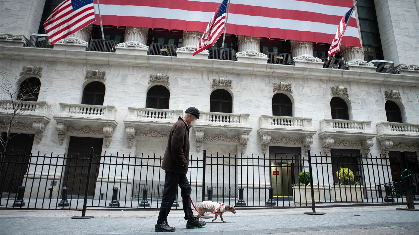 A man in a face mask walking a small dog past the flag-draped front of the New York Stock Exchange