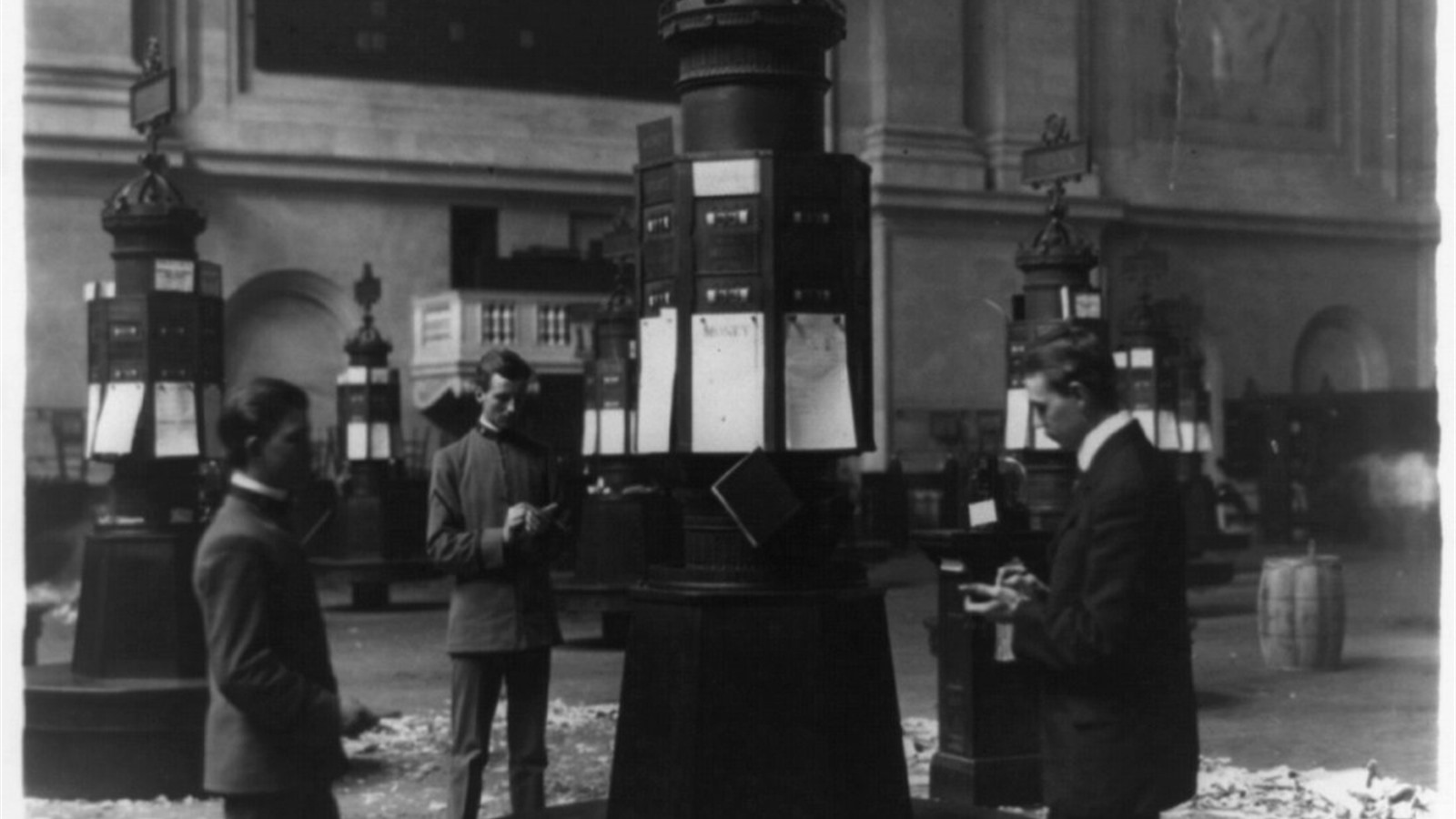 Three men reading share prices at a stock quotations kiosk inside an exchange in 1908